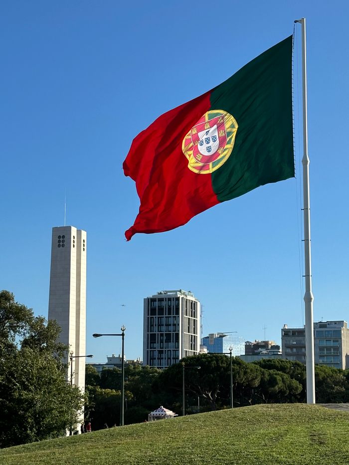 A vibrant Portuguese flag waves prominently over a Lisbon park with a clear urban skyline backdrop.